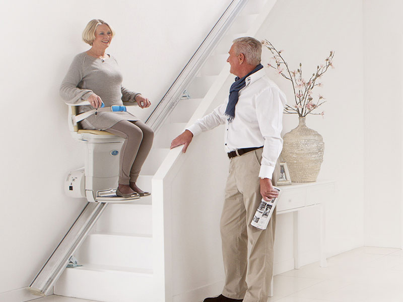 Bild mit einer Frau die auf einem Treppenlift sitzt und die Treppe herauf fährt. Ihr Mann steht unten an der Treppe.